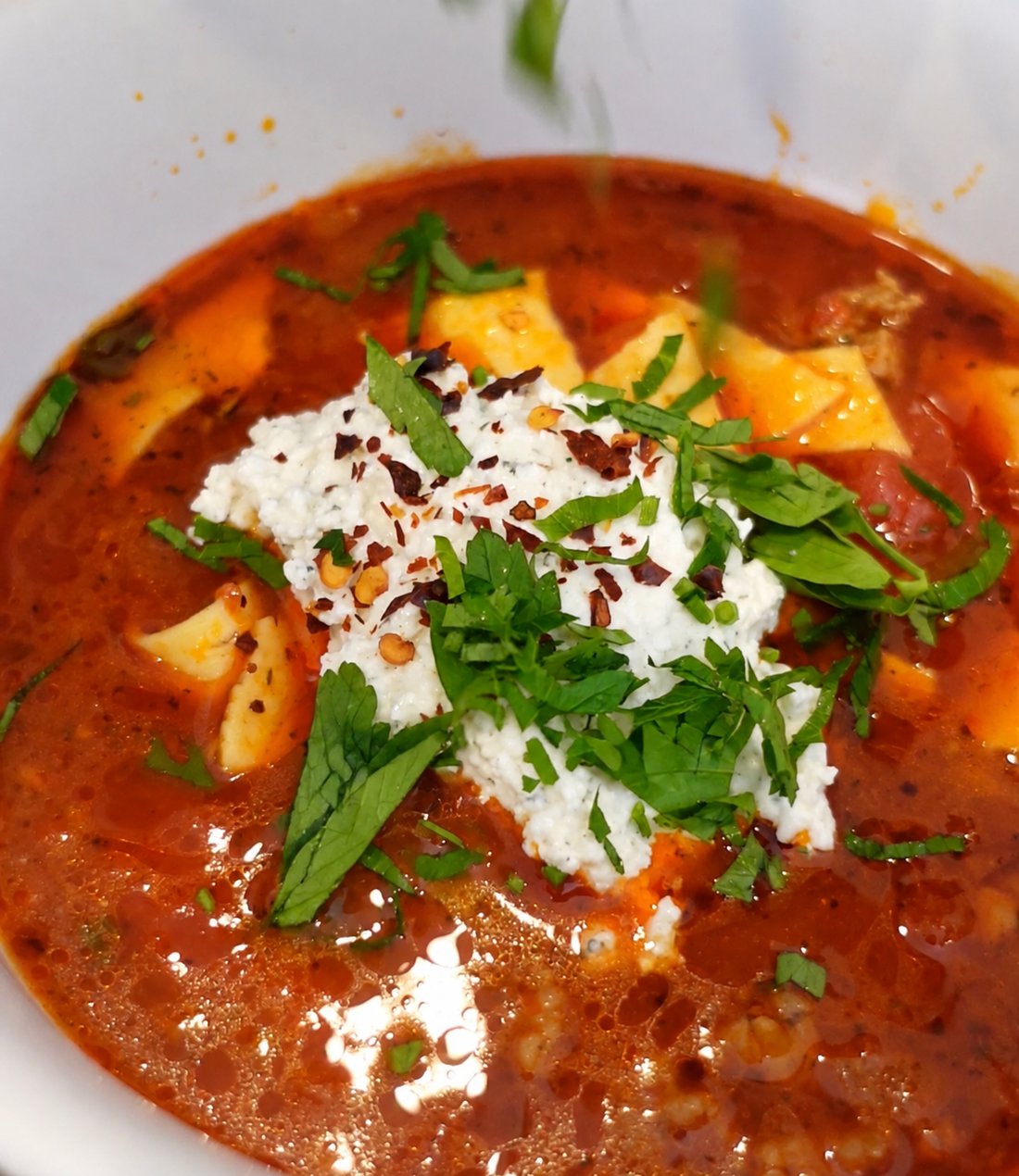 Lasagna soup with broken pasta sheets, whipped ricotta, chopped herbs and chili flakes in a white bowl
