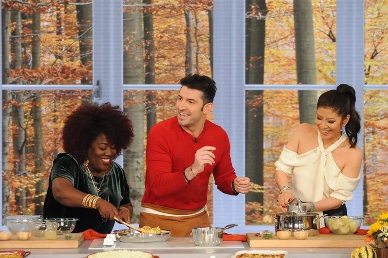 A studio cooking demonstration with three people against an autumn backdrop.