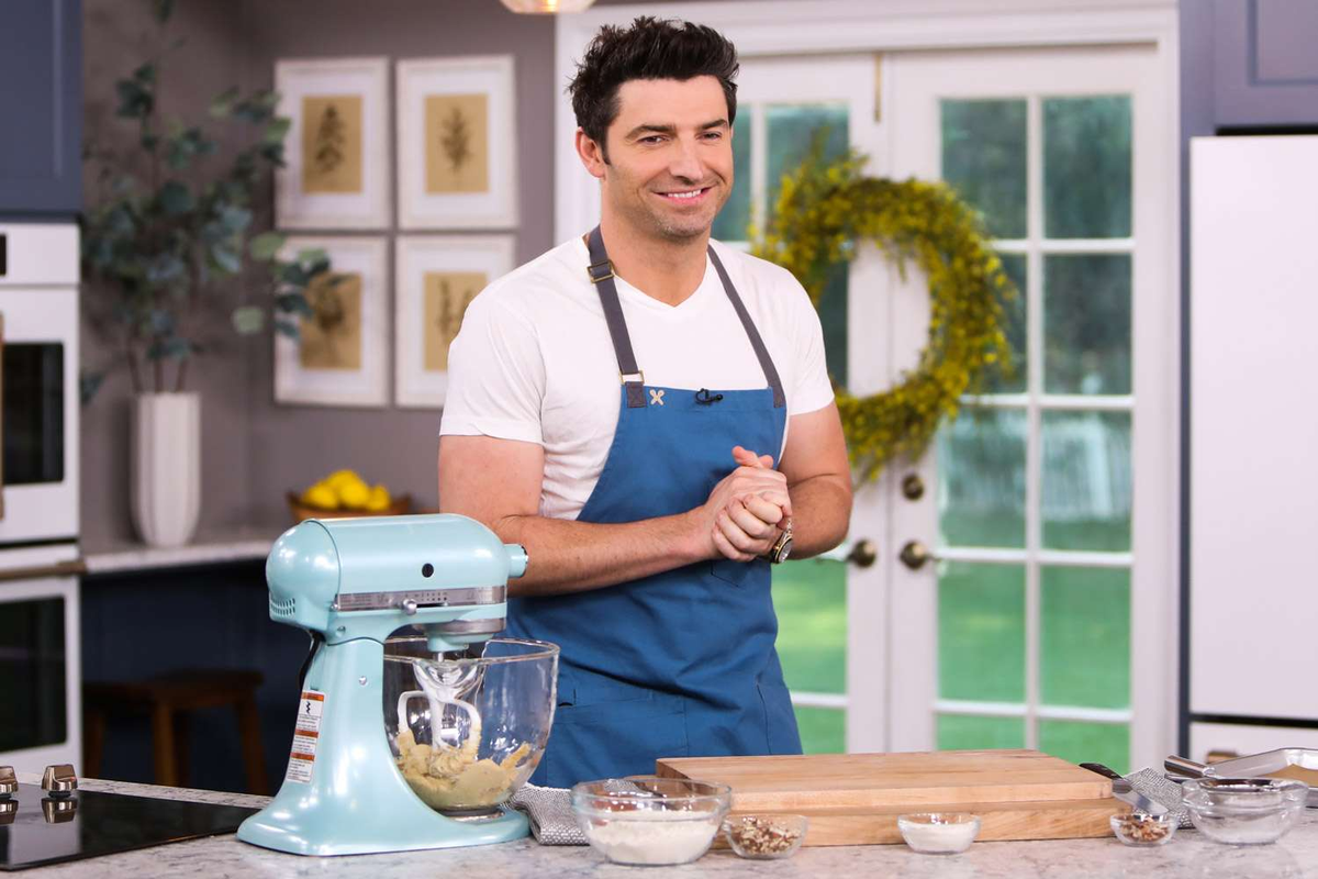 Chef Stuart in a blue apron beside a turquoise stand mixer on a studio kitchen set.