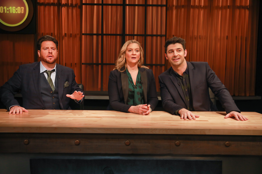 Three judges watch a cooking competition from behind a wooden counter.